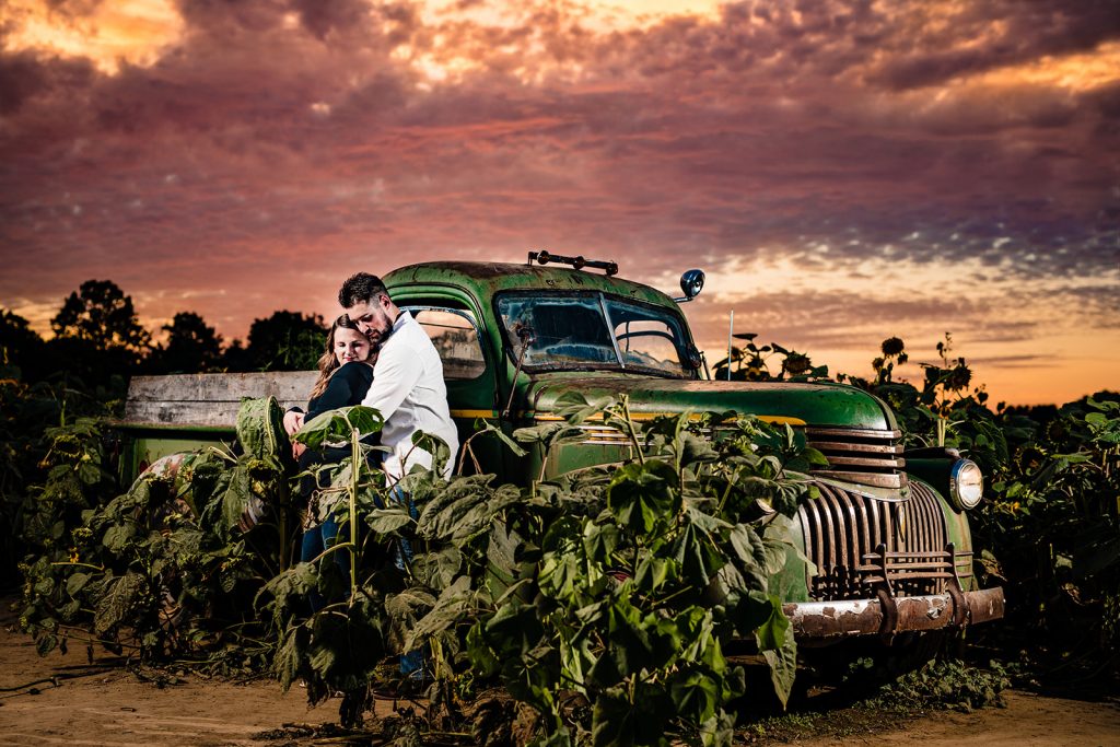 a couple poses by a vintage truck with a gorgeous sunset during their sunflower engagement photos