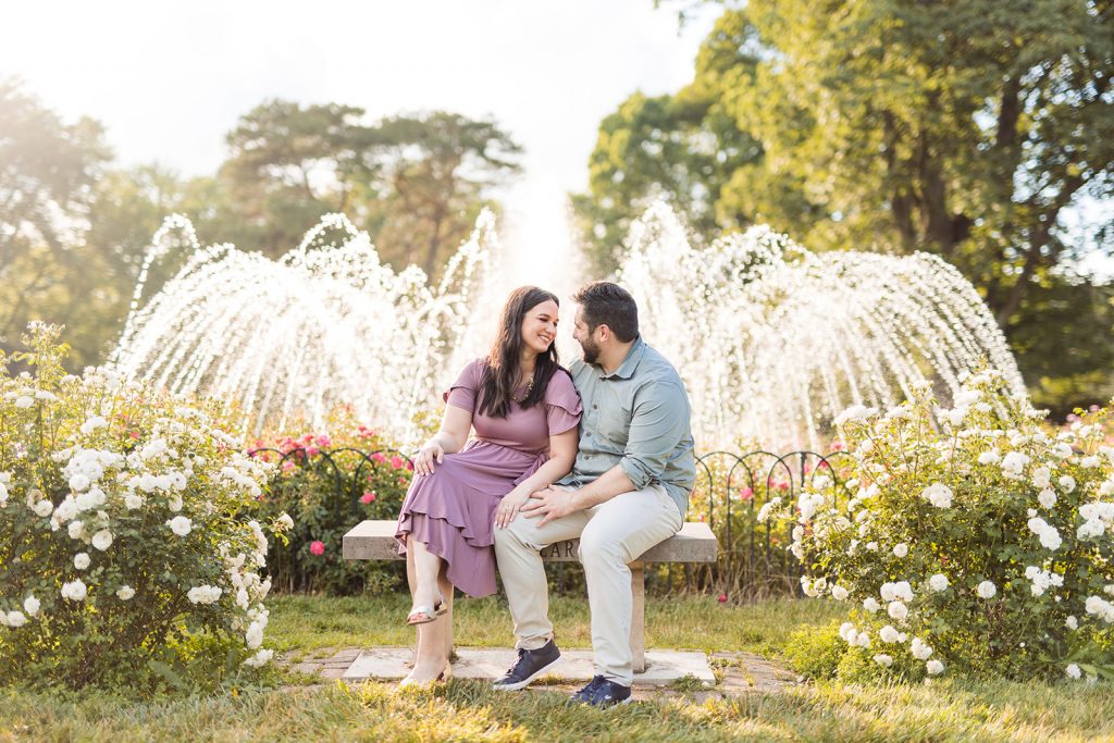 a couple cuddles on a bench in front of a water fountain during their engagement photos at Park of Roses