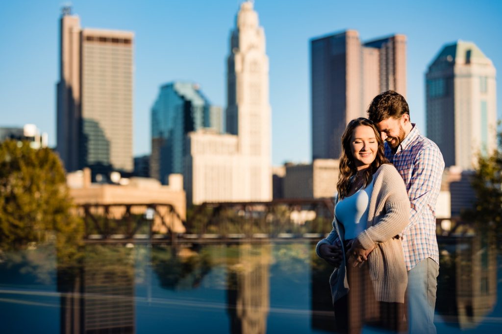 a couple stands in front of the Columbus skyline during their engagement session at North Bank Park