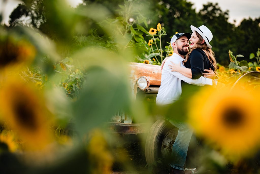 a couple sit on a vintage tractor and laugh during their sunflower engagement photos
