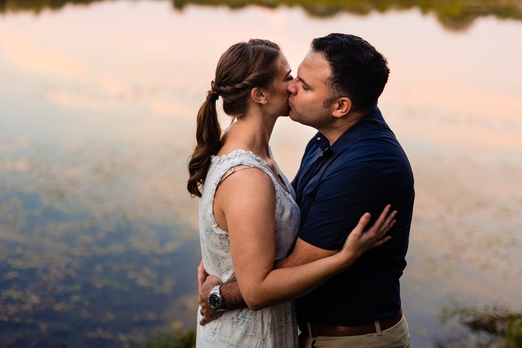 a couple kisses in front of the lake with a sunset reflected in it during their engagement photos at Prairie Oaks Metro Park